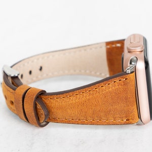 May include: A brown leather watch strap with a rose gold watch face. The strap is stitched with brown thread and has a buckle fastening.