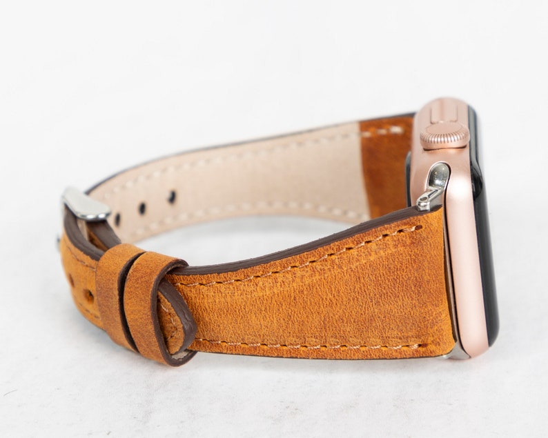 May include: A brown leather watch strap with a rose gold watch face. The strap is stitched with brown thread and has a buckle fastening.
