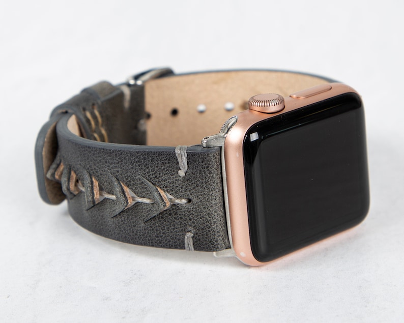 May include: A grey leather Apple Watch band with a rose gold watch face. The band has a stitched design with a chevron pattern.