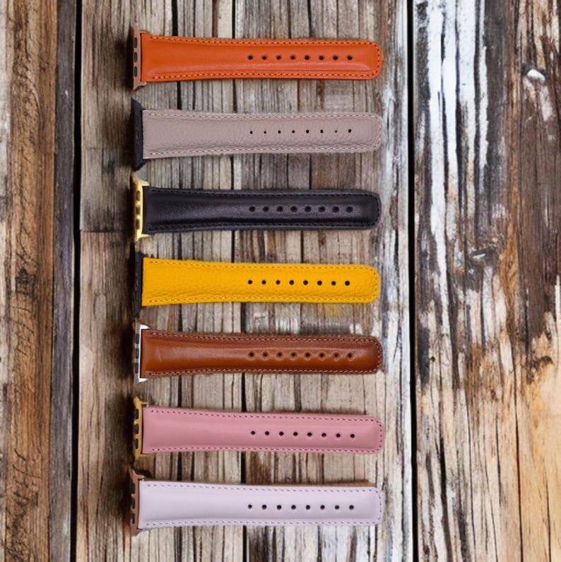 K&ouml;nnte beinhalten: Eine Auswahl an Leder-Armb&auml;ndern in verschiedenen Farben, darunter Orange, Taupe, Schwarz, Gelb, Braun, Rosa und Lavendel. Jedes Armband hat eine Reihe von L&ouml;chern zur Anpassung und wird auf einer Holzoberfl&auml;che pr&auml;sentiert.