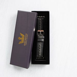 The watch band and rose gold watch case displayed inside an open dark purple gift box with gold logo lettering.