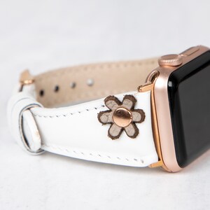 Pode incluir: Pulseira de rel&oacute;gio Apple de couro branco com fivela de ouro rosa e um pequeno detalhe de flor marrom.