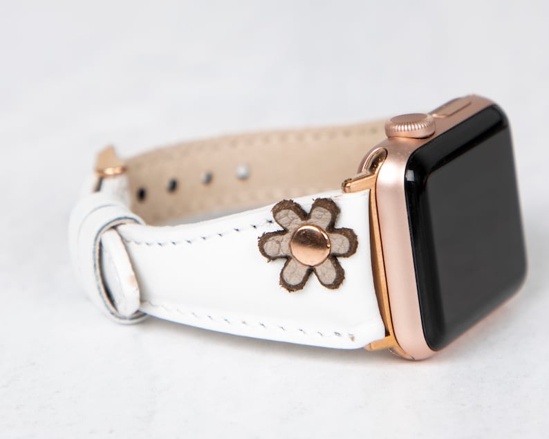 Pode incluir: Pulseira de rel&oacute;gio Apple de couro branco com fivela de ouro rosa e um pequeno detalhe de flor marrom.