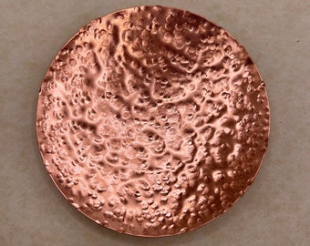 May include: A round copper tray with a hammered texture. The tray has a slightly raised edge and a smooth, polished finish.