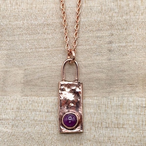 May include: A copper-toned metal pendant necklace featuring a rectangular, hammered-texture plate with a small, circular, deep purple cabochon stone set near the bottom. The pendant hangs from a matching copper-toned metal link chain.