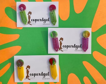 May include: Three pairs of colorful pencil-shaped earrings with a leopard print design. The earrings are displayed on white cards with the text "leopardgal design".