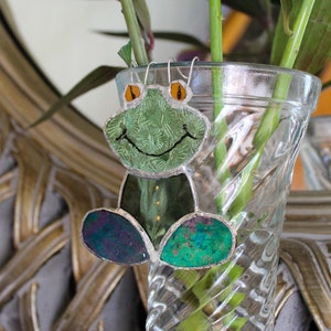 May include: A green frog-shaped stained glass ornament with orange eyes hangs from a clear glass vase filled with green stems.
