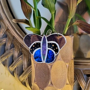 May include: A gold vase with a textured pattern holds a bouquet of green and purple lilies. A stained glass butterfly with blue, purple, and black accents is attached to the vase.