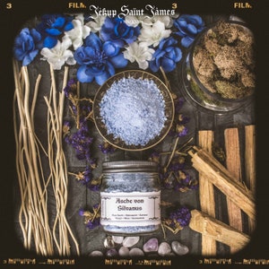 May include: A glass jar filled with blue bath salts labeled "Asche von Silvanus" with a list of ingredients including Palo Santo, Bergamot, Saffron, Violet, Moss, and Sandalwood. The jar is surrounded by dried flowers, wooden sticks, and small purple and pink stones.