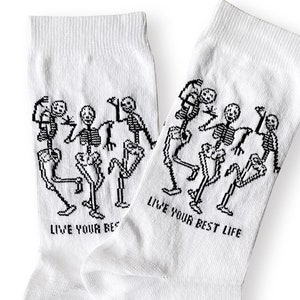 May include: A pair of white socks with a black graphic of dancing skeletons. The text "LIVE YOUR BEST LIFE" is printed in black on each sock. The socks are made of a soft, comfortable material.