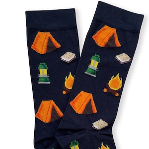 Può includere: Un paio di calzini blu navy con un motivo ripetuto di tende da campeggio arancioni, lanterne verdi, falò gialli e s'mores. I calzini presentano un bordo a coste nella parte superiore e sono disposti sovrapposti su uno sfondo a tinta unita.