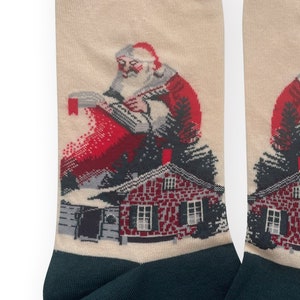 May include: A pair of cream-colored socks with a green cuff. The socks feature a vintage illustration of Santa Claus delivering presents to a house with snow on the roof.