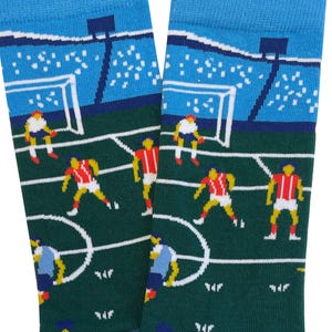 Op de afbeelding: Een paar sokken met een voetbalveldontwerp. De sokken hebben een groen veld met witte lijnen, spelers in rode en blauwe uniformen en een stadionachtergrond in blauw en wit.