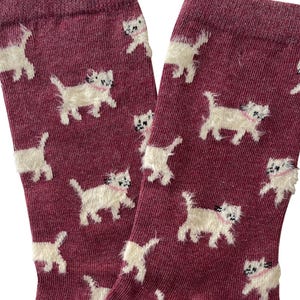 May include: Burgundy socks featuring a repeating pattern of white, fluffy cat illustrations, each with a pink collar. The socks are made from a soft, textured material.