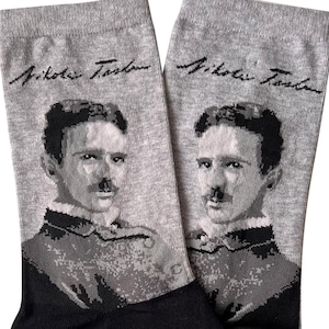 Könnte beinhalten: Ein Paar graue Socken mit einem Schwarzweißbild von Nikola Tesla auf jeder Socke. Die Socken haben den Text "Nikola Tesla" in Schwarz auf der Oberseite jeder Socke.