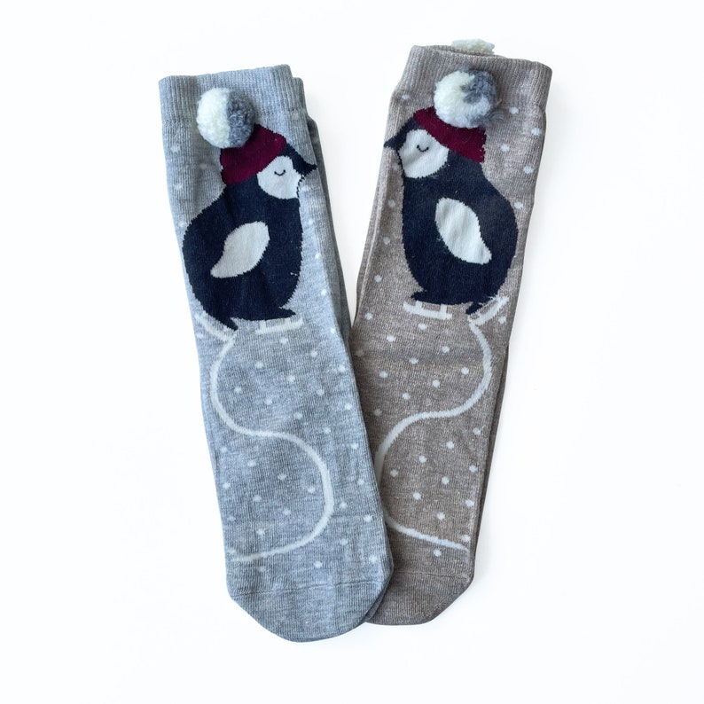 May include: Two pairs of knee-high socks. One pair is light grey, the other is tan. Each sock features a penguin wearing a red hat with a white pom-pom, ice skating on a white dotted background. The socks are designed for children.