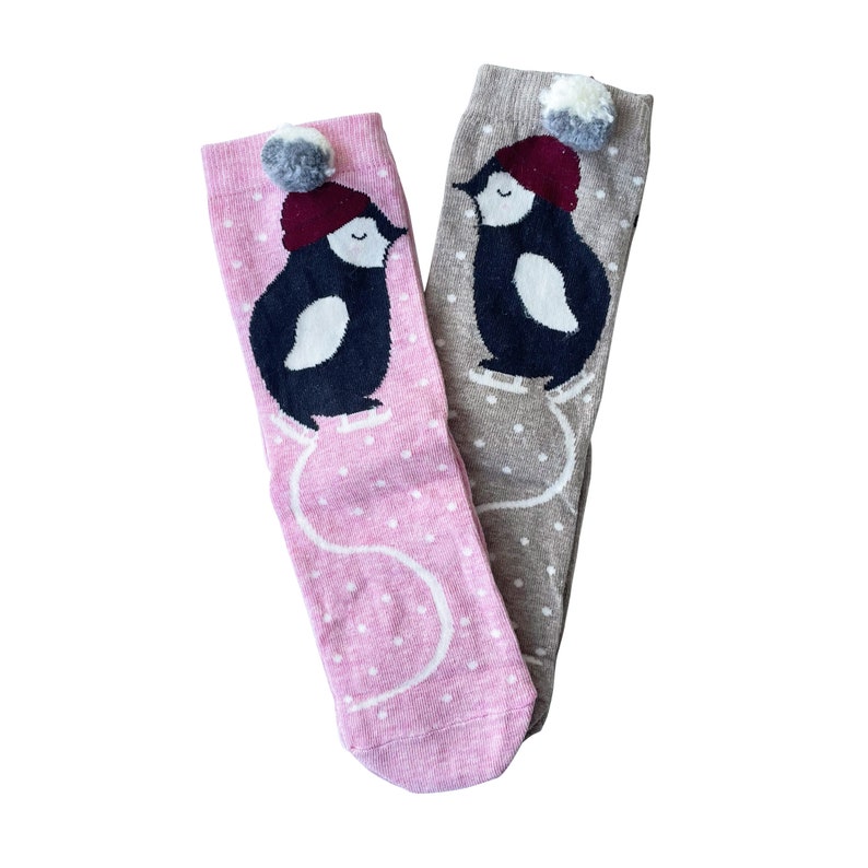 May include: Two pairs of knee-high socks. One pair is pink with white polka dots, the other is grey with white polka dots. Each sock features a penguin graphic wearing a red hat with a grey pom-pom. The penguins appear to be ice skating.