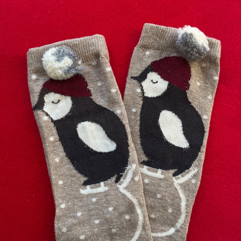 May include: A pair of tan socks with a penguin design. The penguins are black and white with burgundy hats and white ice skates. Each sock has a grey and white pom-pom at the top and white polka dots. The socks are on a red background.