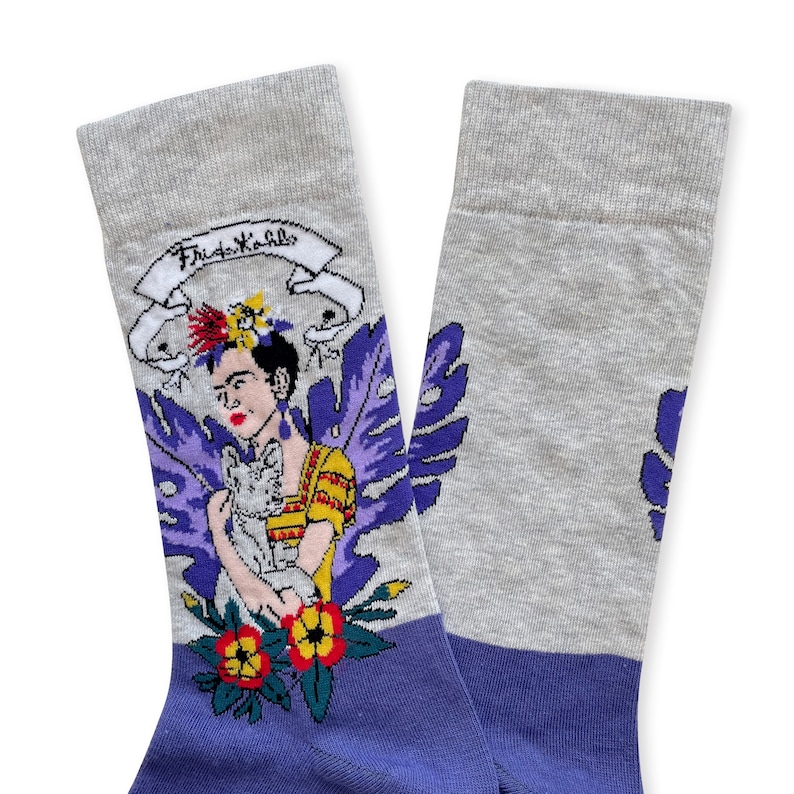 May include: A pair of grey and purple socks with a colorful illustration of Frida Kahlo holding a cat. The socks have the text "Frida Kahlo" on the top of the sock.