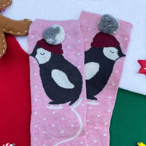 May include: A pair of pink socks with a penguin design wearing a burgundy hat and a grey and white pom-pom. The penguin is black and white, with white polka dots on the socks. A festive holiday item.