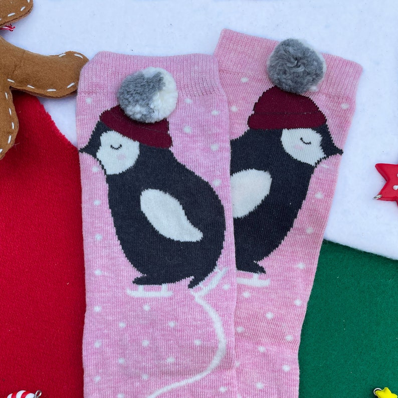 May include: A pair of pink socks with a penguin design wearing a burgundy hat and a grey and white pom-pom. The penguin is black and white, with white polka dots on the socks. A festive holiday item.