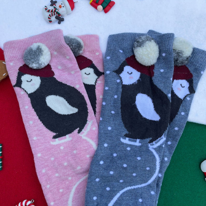May include: Pink and grey socks featuring a penguin design. The penguins wear red hats and ice skates, topped with a white pom-pom. White polka dots are scattered across the socks. A festive winter accessory.