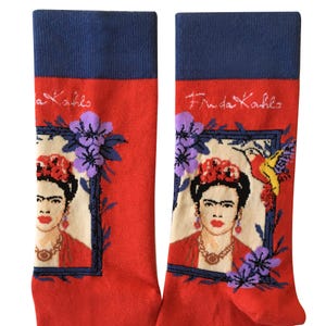May include: A pair of red socks with a blue cuff. The socks feature a portrait of Frida Kahlo in a frame with flowers and a hummingbird. The text "Frida Kahlo" is written on the cuff of the socks.