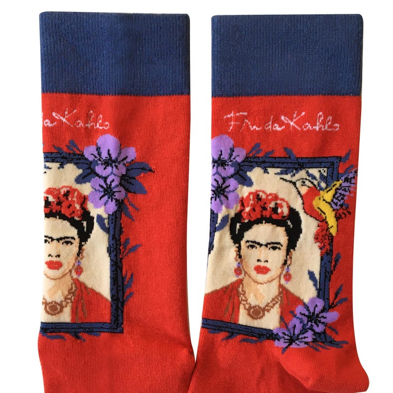 May include: A pair of red socks with a blue cuff. The socks feature a portrait of Frida Kahlo in a frame with flowers and a hummingbird. The text "Frida Kahlo" is written on the cuff of the socks.