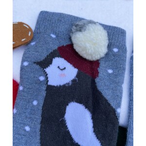 May include: Close-up of a grey sock featuring a penguin design. The penguin has a white belly, a dark grey body, and a red hat. A white pom-pom is attached to the top of the sock. White snowflakes are scattered across the grey background.