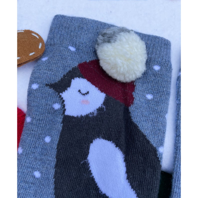 May include: Close-up of a grey sock featuring a penguin design. The penguin has a white belly, a dark grey body, and a red hat. A white pom-pom is attached to the top of the sock. White snowflakes are scattered across the grey background.