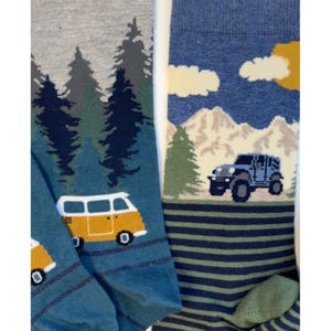 May include: Two pairs of socks with outdoor-themed designs. One pair features a yellow camper van in front of a forest scene. The other pair has a blue jeep in front of mountains and trees. Both pairs have blue and green striped patterns.
