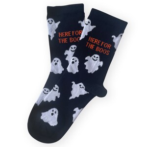 May include: A pair of black socks with white ghost illustrations. The socks have the text "HERE FOR THE BOOS" in orange. The ghosts have smiling faces. These socks are perfect for Halloween.