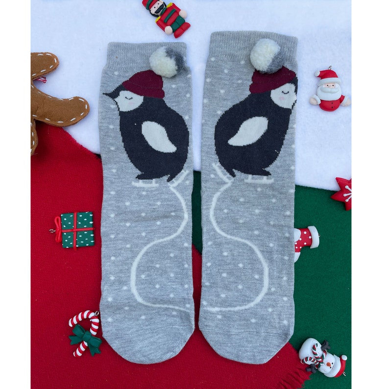 May include: A pair of grey socks with a penguin design. Each sock features a black penguin wearing a burgundy hat and white polka dots. The socks have a white pom-pom and are decorated with Christmas-themed accessories.