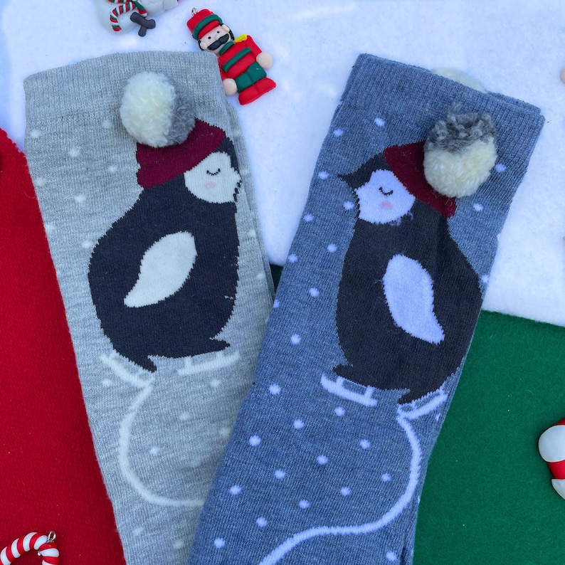 May include: Two pairs of knee-high socks featuring a penguin design. The penguins wear red hats and ice skate on white skates. The socks are grey and blue with white polka dots and a white pom-pom.