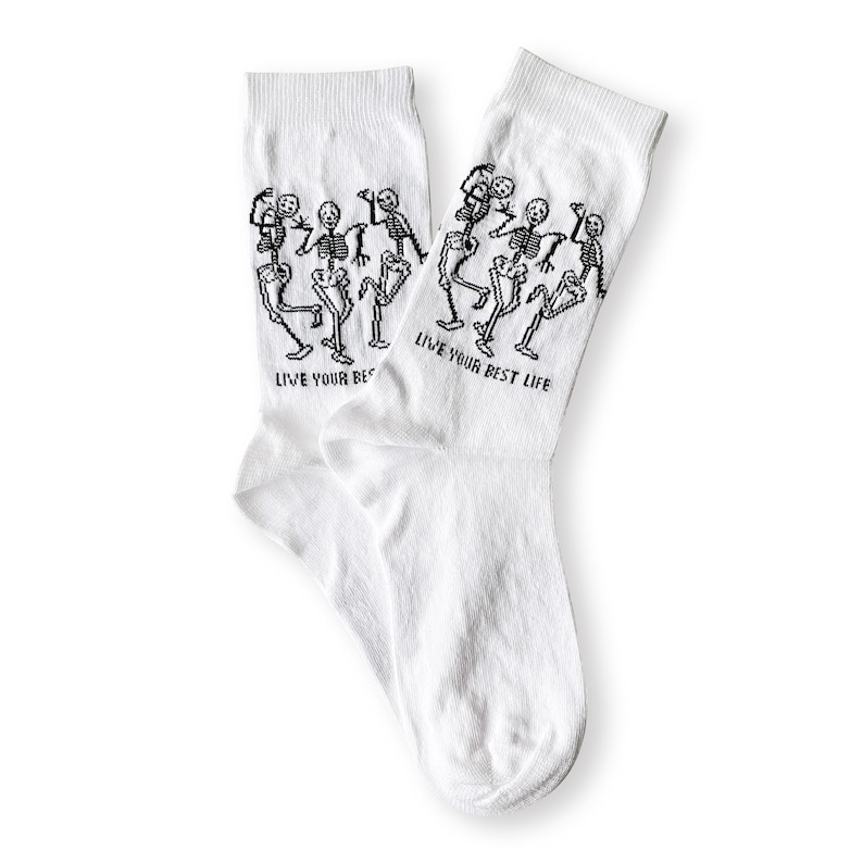 May include: A pair of white socks with a black graphic of dancing skeletons. The text "LIVE YOUR BEST LIFE" is printed in black on each sock. The socks are made of a soft, comfortable material.
