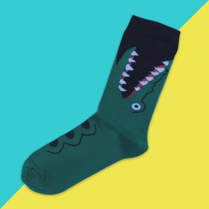 May include: Green and black socks featuring a cartoon alligator design. The alligator has a red and white striped mouth and a single eye.
