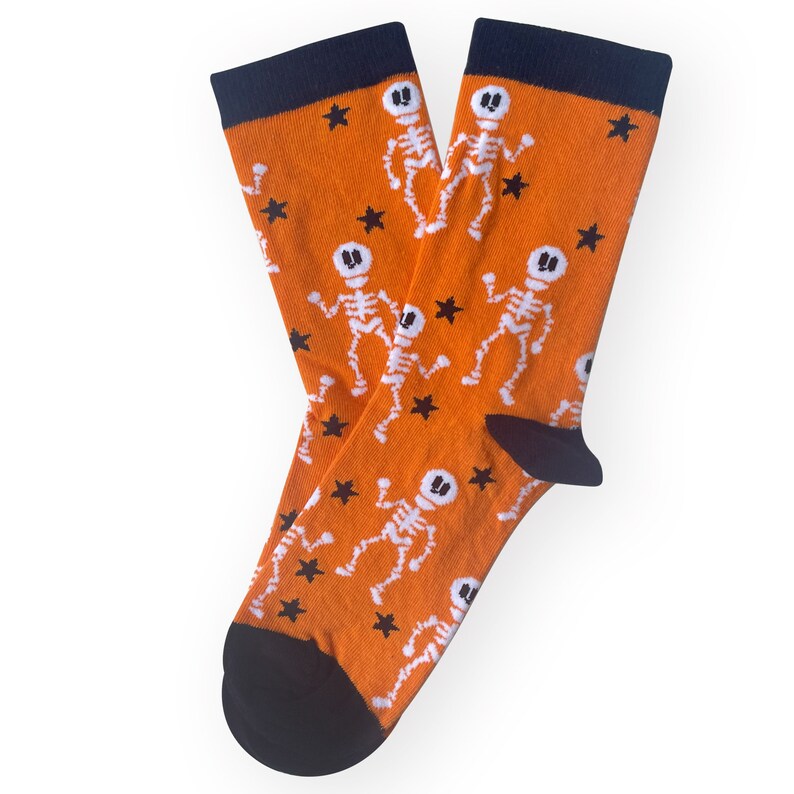 May include: A pair of orange socks with a navy blue cuff and toe. The socks feature a repeating pattern of white skeleton figures and black stars, perfect for Halloween.