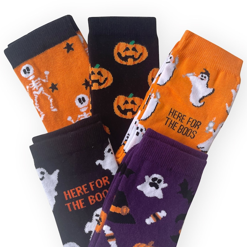May include: Five pairs of Halloween-themed socks in various colors. The socks feature designs of skeletons, jack-o'-lanterns, ghosts, bats, and witch hats. Some socks have the text "HERE FOR THE BOOS". Colors include orange, black, and purple.