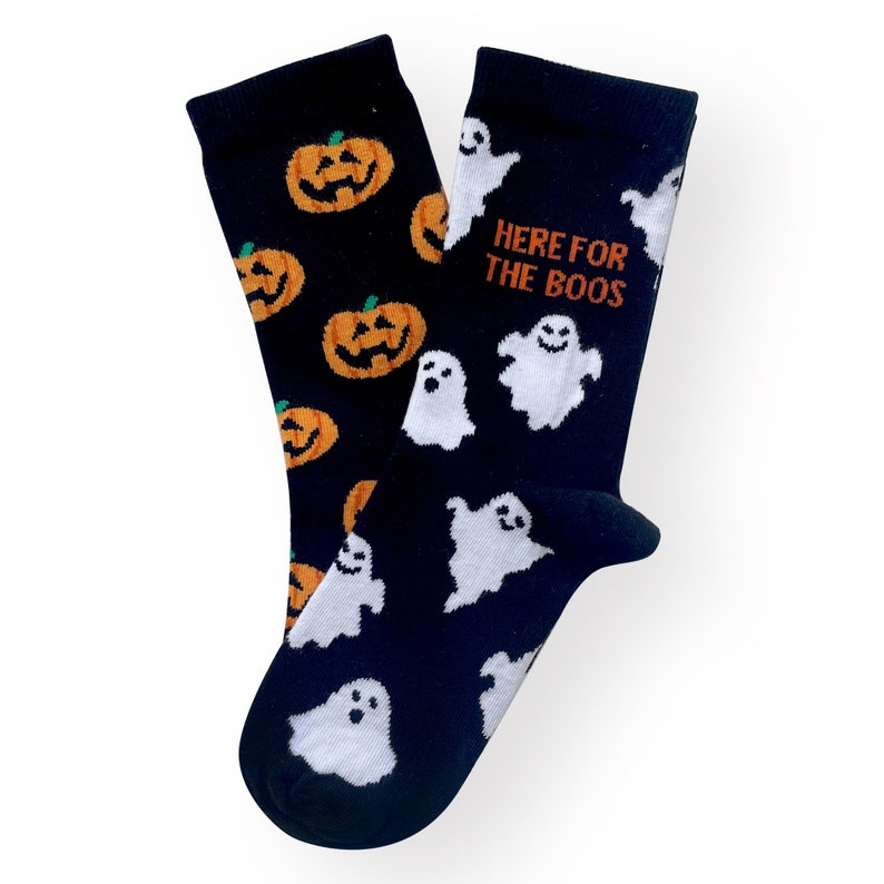 May include: A pair of Halloween-themed socks. One sock features orange jack-o'-lanterns on a black background. The other sock has white ghosts and the text "HERE FOR THE BOOS" on a black background. The socks are made of a soft material.