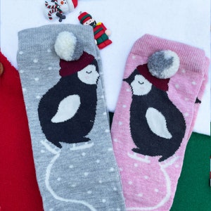 May include: Two knee-high socks, grey and pink, with penguin designs. The penguins wear burgundy hats and skate on white lines. Each sock has a pom-pom and white polka dots. Christmas ornaments are in the background.