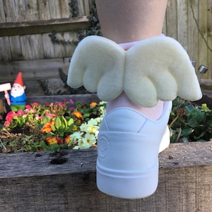 Può includere: Scarpe da ginnastica bianche con un accessorio ad ali d'angelo giallo chiaro. Le ali sono attaccate a un calzino rosa. Le scarpe da ginnastica sono su una superficie di legno con un giardino sullo sfondo.