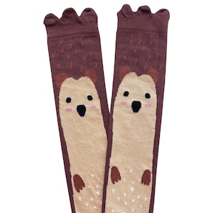 Peut inclure: Une paire de chaussettes mi-hautes brunes et beiges avec un motif de hérisson. Les chaussettes ont un haut festonné et un visage de hérisson mignon avec des joues roses et des yeux noirs.