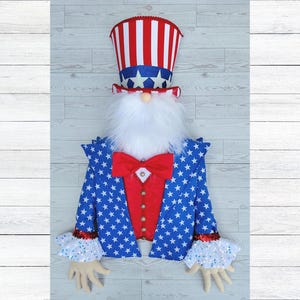 May include: A patriotic Uncle Sam gnome decoration. The gnome features a red, white, and blue top hat, a white beard, and a blue jacket with white stars. A red bow tie and a red vest with gold buttons complete the festive look.