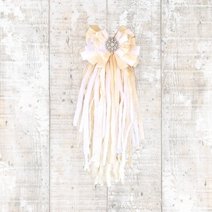 May include: A decorative bow with a rhinestone center and long, flowing fabric strips. The bow features layers of cream and pale yellow fabric, creating a textured and elegant design. The background is a light wood-grain pattern.