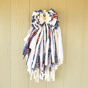 May include: A patriotic wreath with a large bow and cascading fabric strips. The bow features layers of white, red, and blue fabric with star and stripe patterns. A central flower with a rhinestone adds a touch of elegance. The wreath is designed for door or wall decor.