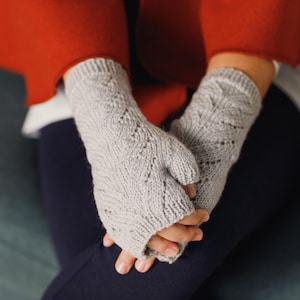 May include: Light grey fingerless gloves with a detailed knitted design. The gloves are made of soft yarn and have a thumb opening. The person is wearing a red jacket and dark blue trousers.