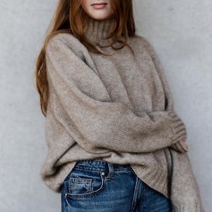May include: A beige roll-neck jumper with long sleeves. The jumper is worn with blue denim jeans. The jumper has a ribbed texture on the cuffs and hem. The model is leaning against a grey wall.
