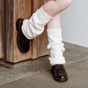 May include: Off-white leg warmers worn with black leather shoes. The leg warmers are a chunky knit design, gathered at the ankles. The shoes are lace-up with a rounded toe and a thick sole. The background features a wooden door and a concrete floor.