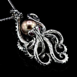 May include: A silver octopus pendant with a large, brown pearl in the center. The octopus's tentacles are curled around the pearl, and there are small red gemstones on the octopus's head.