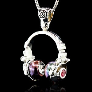 May include: A silver pendant necklace featuring a detailed headphone design. The headphones have iridescent pearl-like earcups and a small red gemstone on one side. The pendant hangs from a delicate silver chain against a black background.